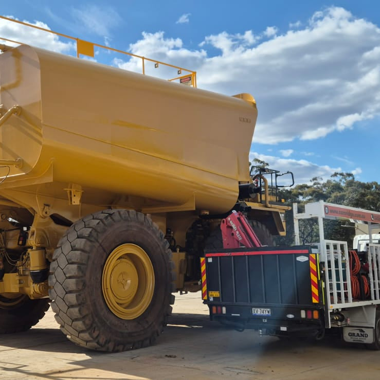 A large yellow mining haul truck with oversized tires parked outdoors under a blue sky, next to a white shipping container and a utility vehicle.