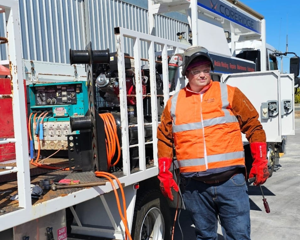 A worker in a high-visibility orange safety vest and blue jeans stands beside a white service truck outdoors, with orange extension cords coiled on the ground under a clear blue sky.
